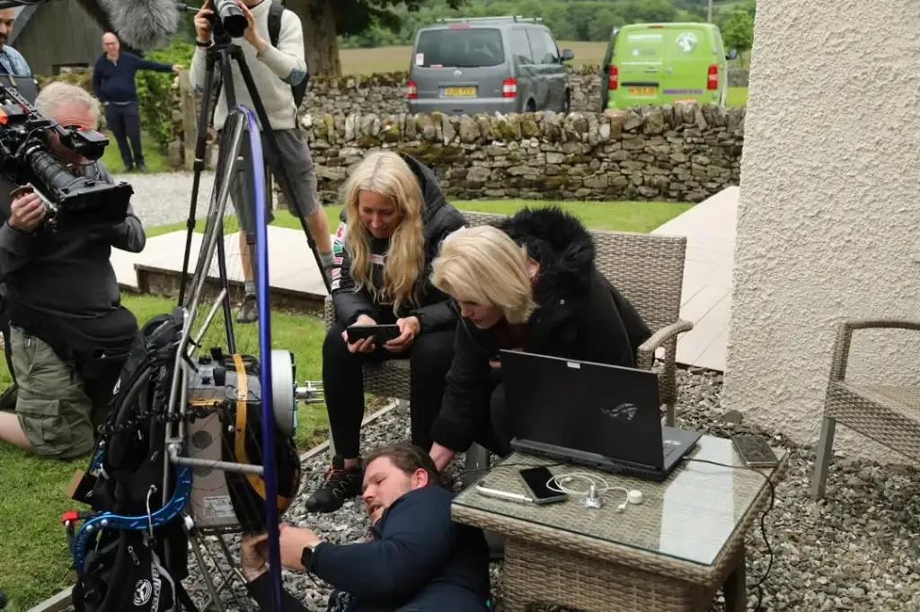 An ITV camera operator films outdoors while two women look at a phone and a laptop at a garden table, and Louis Wright lies on the gravel adjusting equipment on a tripod.